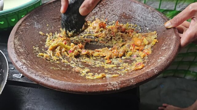 Person using a traditional stone pestle to grind chilies and shallots into a spicy paste on a flat mortar