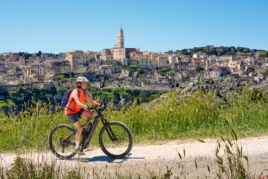 Active woman riding an electric mountain bike with a panoramic view of the medieval cave city skylineof Matera, Basilicata, Italy
