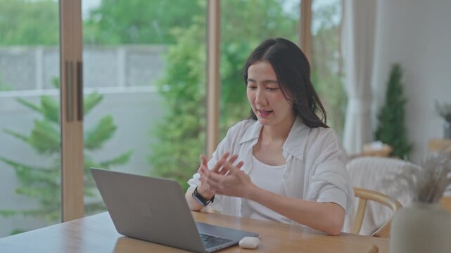 Focused young Asian woman gesturing with hands while talking on a video call using a laptop, working remotely at a wooden table in a bright modern home with a garden view.