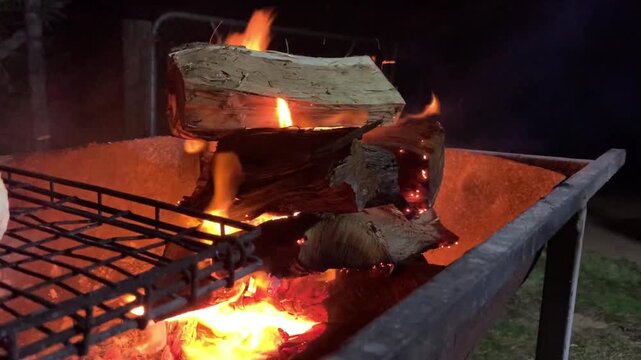 Logs burning in a metal barbecue grill at night with glowing embers