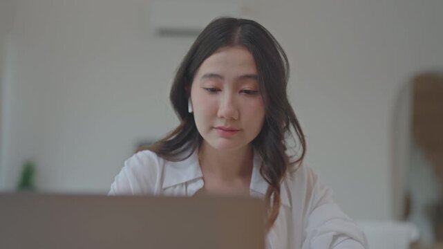Close up portrait of Asian woman with wireless earbuds focusing on laptop screen, working and searching for information online at home in a bright minimal room