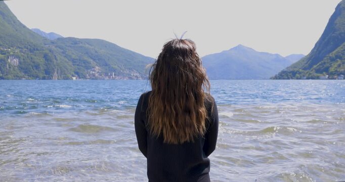A woman gazes at the distant horizon in front of her in front of Lake Ceresio in Lugano, Switzerland, with mountains and hills forming the backdrop, and a light breeze blowing, making the model's long