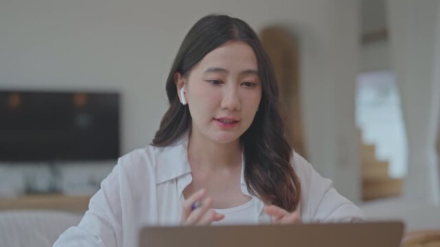 Asian woman wearing wireless earbuds speaking with an AI voice assistant on laptop, using chatbot technology to work from home, talking and listening in a bright room