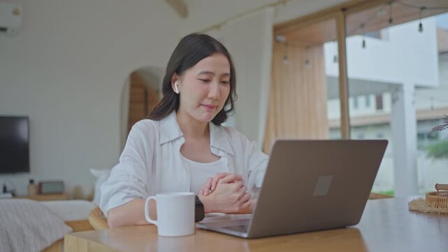 Asian woman wearing earbuds speaking with AI voice assistant on laptop at home, using chatbot for online learning and e-learning, talking and studying with technology