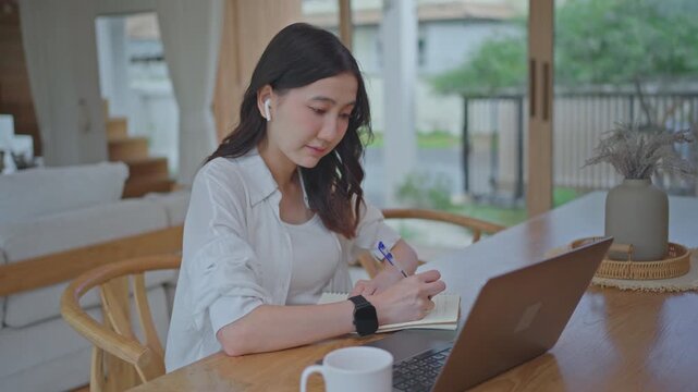 Asian woman wearing earbuds typing on laptop with deep focus, working from home and concentrating on her tasks at a wooden table in a bright modern room