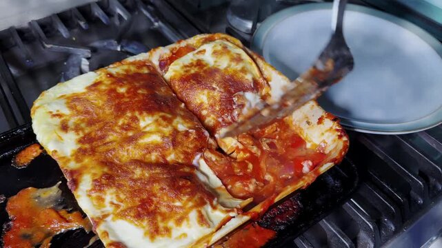 Closeup of a woman&rsquo;s hand using a slice tool to cut and lift a portion of steaming mixed vegetable lasagne, from a baking dish, ready to place on a nearby plate.