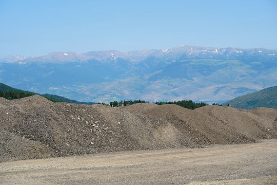 A wide shot at a road construction site showing a row of large gravel and crushed stone mounds on the ground that mimic the shapes of majestic mountain ranges with snow peaks in the distant background