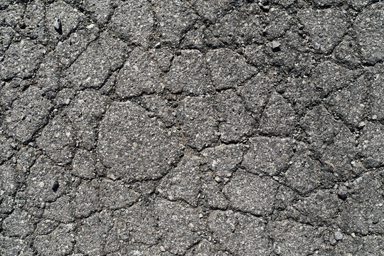 A full-frame top-down close-up shot of heavily cracked, broken, and damaged old grey asphalt pavement. The deep network of fissures symbolizes infrastructure decay and the need for road repair.