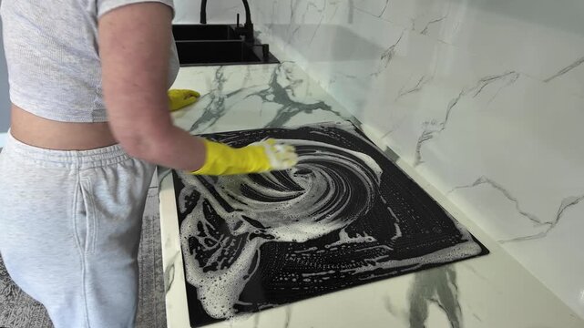 Woman wearing yellow rubber gloves cleaning a black induction cooktop. Professional housekeeper wiping the stove with a foamy sponge