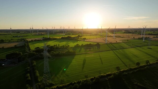 Aerial footage of wind farm and high voltage power lines at sunset over agricultural landscape. Renewable energy infrastructure for the energy transition.