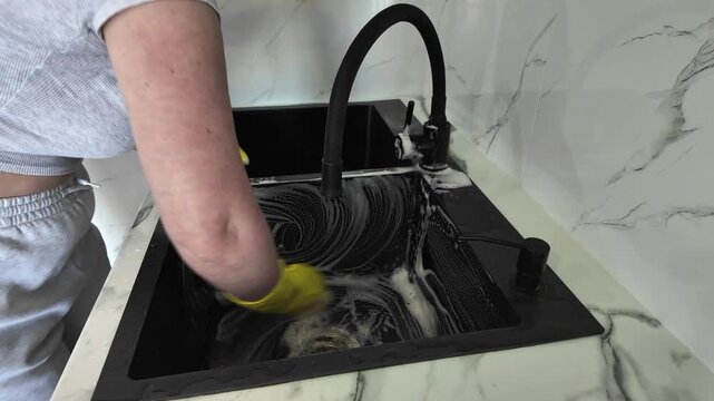 Woman in yellow rubber gloves diligently scrubbing a modern black kitchen sink with a sponge and soapy foam. Household chores and hygiene concept