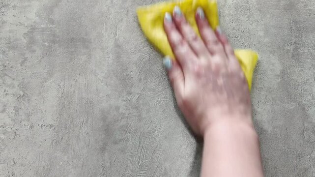 A woman's hand wipes the textured gray surface of a table with a yellow rag