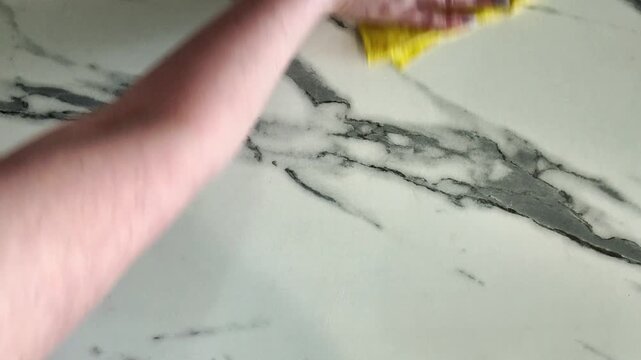 Female hand using a yellow microfiber cloth to wipe and disinfect a white marble kitchen countertop. Routine household chore for home hygiene