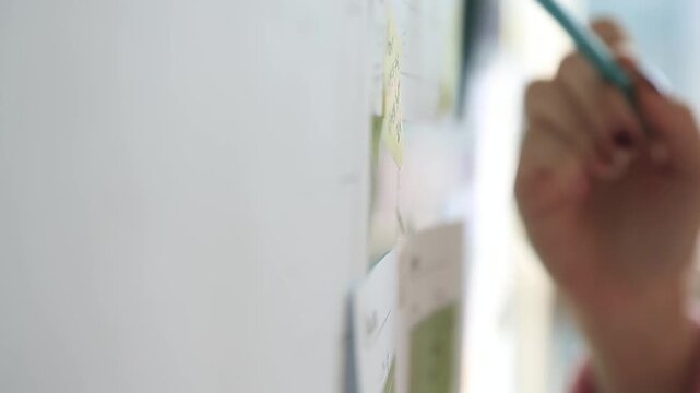 사람 일상 소품 디테일 자재: 실내 사무실 보드판에 붙은 노란색 메모지에 하늘색 필기구를 대고 무언가를 가리키거나 표시하는 사람의 손동작을 가까이서 담은 클로즈업 동영상 클립입니다.