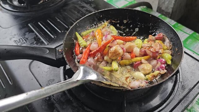 Person stirring sizzling shallots garlic and fresh chilies in hot oil with a metal spatula on a gas stove making a spicy condiment