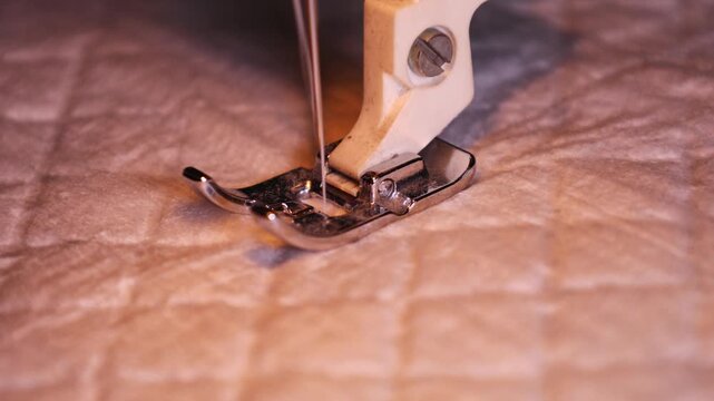 Sewing machine needle piercing soft quilted fabric, performing precise stitching. Focused view of metal presser foot and needle combined with textile texture, illuminated by warm light.
