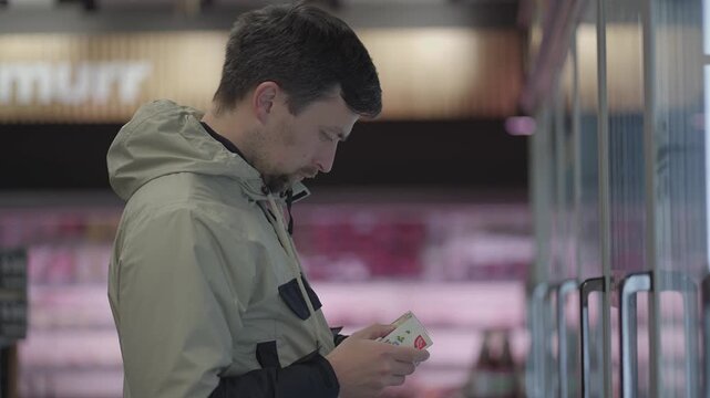 A male shopper carefully examines the ingredients and nutrition facts on a package of frozen ready meal after taking it from a commercial supermarket freezer display with a glass door.