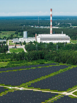 Aerial view of a large solar farm with rows of photovoltaic panels situated near a power plant with a tall chimney surrounded by green forests in Riga, Latvia.