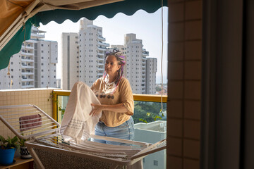 A woman with dreadlocks taking a white towel from a drying rack on a balcony with city high-rise buildings in the background. © elenarostunova
