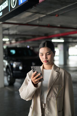 Fototapeta premium Young Thai Isan Woman Using Phone next to Black Car in Parking Garage with Copy Space