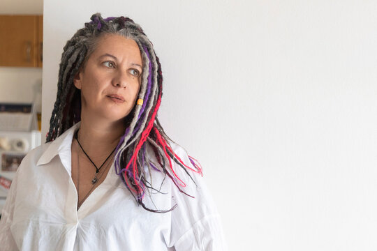 Portrait of a mature woman with colourful dreadlocks wearing a white shirt looking aside indoors.