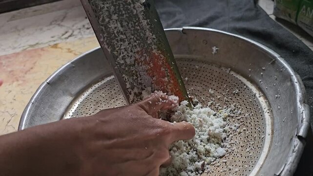 A person manually grating fresh coconut meat using a traditional metal grater over a strainer for cooking preparation