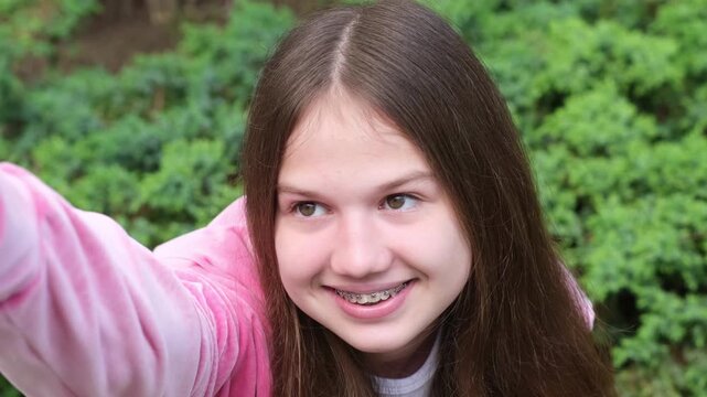 Happy smiling teenage girl with dental braces taking selfie on green nature background outdoors. Teeth health care, orthodontic concept
