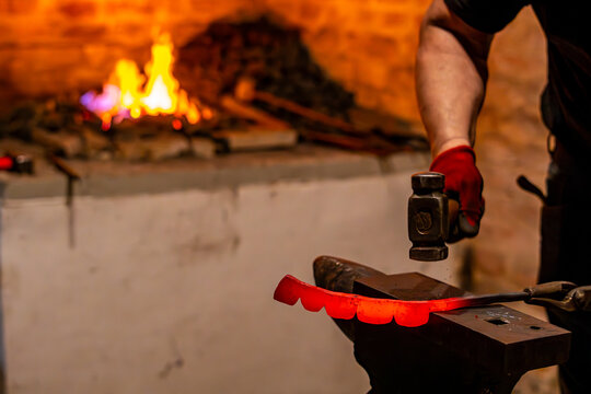 Blacksmith hammering a glowing red-hot metal piece on an anvil, forge furnace with bright fire in the background, traditional craftwork inside a dark workshop.