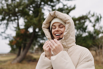 Woman in a beige winter coat and hood smiling warmly outdoors on a cold day, enjoying nature, fresh air, and subtle sunlight. © SHOTPRIME STUDIO