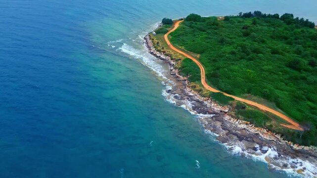 An aerial view captures a rugged coastline with waves crashing against rocky shores. A lone vehicle traverses a winding dirt road, bordered by lush greenery.