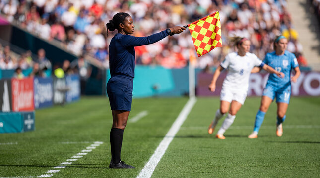 Professional female soccer referee raising a checkered offside flag on the sideline during a women's football match, featuring blurred players and stadium crowd in bright daylight.