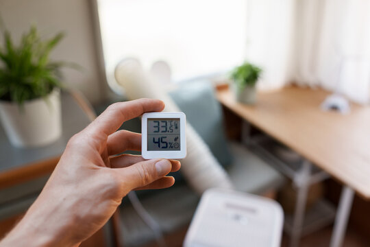 Close-up of a hand holding a room thermometer showing temperature in front of a portable air conditioner
