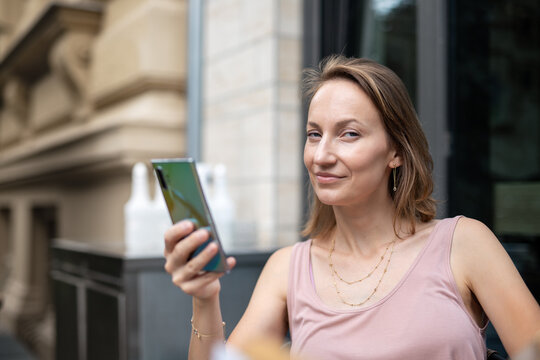 Confident woman uses smartphone while relaxing at outdoor cafe in European city. Modern lifestyle portrait reflects digital communication, remote work, social media, and everyday urban convenience