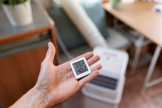Close-up of a hand holding a room thermometer showing temperature in front of a portable air conditioner