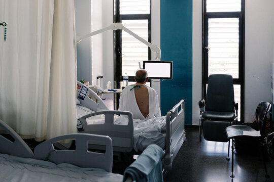 Older patient sits backwards on hospital bed wearing open white gown