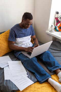 Student working on laptop with documents on bed in dormitory room