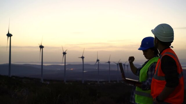 Two workers in safety gear, one wearing a blue hard hat and high-visibility vest, the other in a white hard hat and orange vest, examine wind turbines on a hillside at dusk.