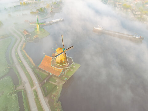 Aerial view of Zaanse Schans windmills surrounded by morning mist over the Zaan river with a cargo ship passing by in Zaandam, Noord-Holland, Netherlands.