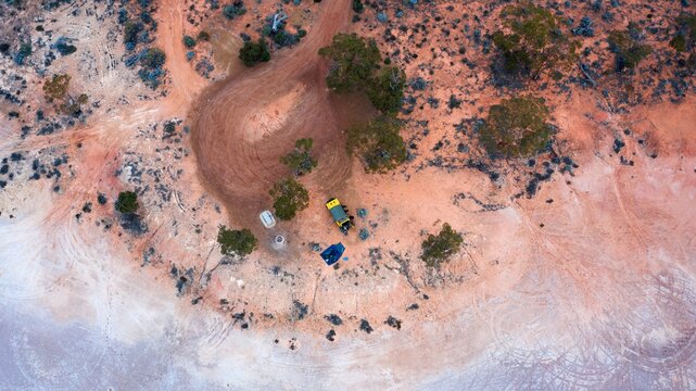 Aerial view of a remote campsite with a yellow vehicle and a blue tent parked on red earth surrounded by sparse eucalyptus trees in the outback near Norseman, Western Australia, Australia.