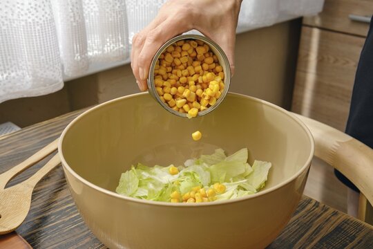 A hand pours canned corn into a bowl filled with fresh green lettuce. The warm kitchen features wood accents, creating a welcoming atmosphere for meal preparation