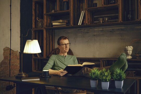 A person sits comfortably in a modern workspace, immersed in a book. Soft lamp light creates a warm atmosphere. Green plants and rustic shelves add character to the relaxing environment