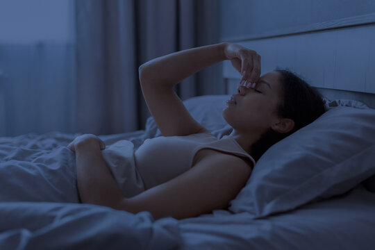 Black woman lying awake in bed at night and pinching her nose, suggesting sinus pressure, headache, insomnia and discomfort in a dark blue bedroom