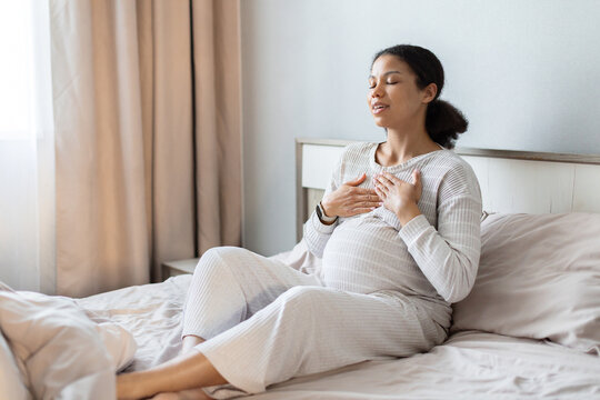 Pregnant black woman sitting on bed and practicing calm breathing with hands on her chest, showing prenatal relaxation and wellness at home