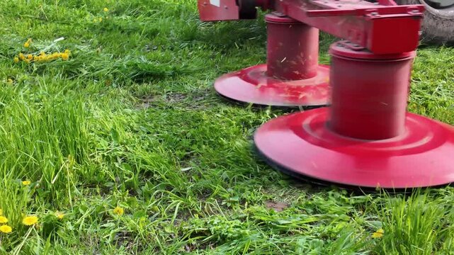 Close-up footage of a rotary mower with spinning drums preparing for hay cutting in a field. The machine is shown in detail as the cutting elements rotate before the tractor begins moving. The tractor