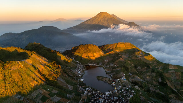 Aerial view of Telaga Cebong lake nestled among terraced agricultural fields and volcanic peaks at sunrise in Indonesia.