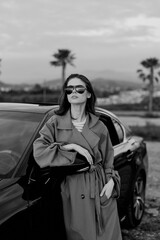 Naklejka premium A stylish woman stands beside a sleek car in a trench coat and sunglasses, exuding confidence. A street lifestyle moment with fashion, independence, and bold attitude.