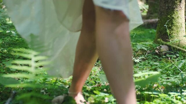 Barefoot woman in white dress walking through forest toward camera, handheld low angle shot