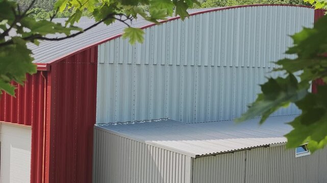Red and grey corrugated metal siding of a modern barn or industrial building. Sunlight casts shadows on the structure, with green leaves framing the view