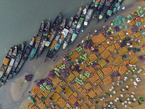 Sylhet, Bangladesh - 07 February 2026: Aerial view of a bustling riverside market with traditional wooden boats docked along the muddy bank, laden with colorful fresh vegetables and produce.