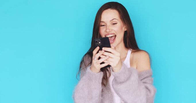 Young woman looking at smartphone screen, celebrating success with ecstatic joyful facial expression, winning gesture, cheering, studio isolated on blue background, concept of victory and excitement